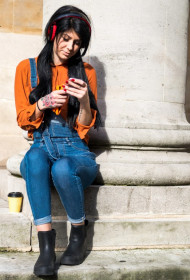 Young woman sitting beside pillar, wearing headphones, holding smartphone