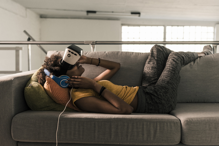 Young woman lying on couch with headphones and VR glasses - Photos