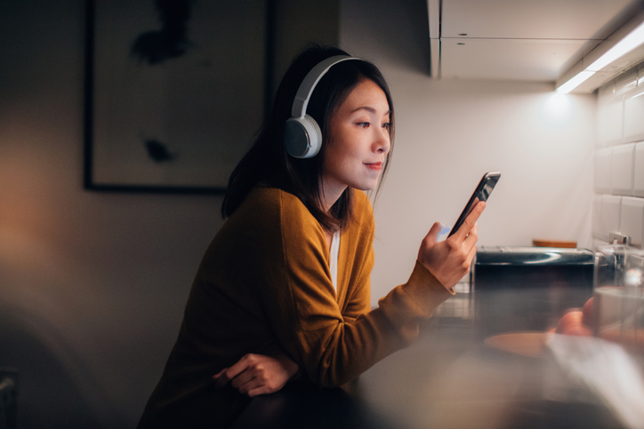 Jeune femme asiatique écoutant un podcast sur son smartphone le soir à la maison. Le téléphone comme outil de divertissement.