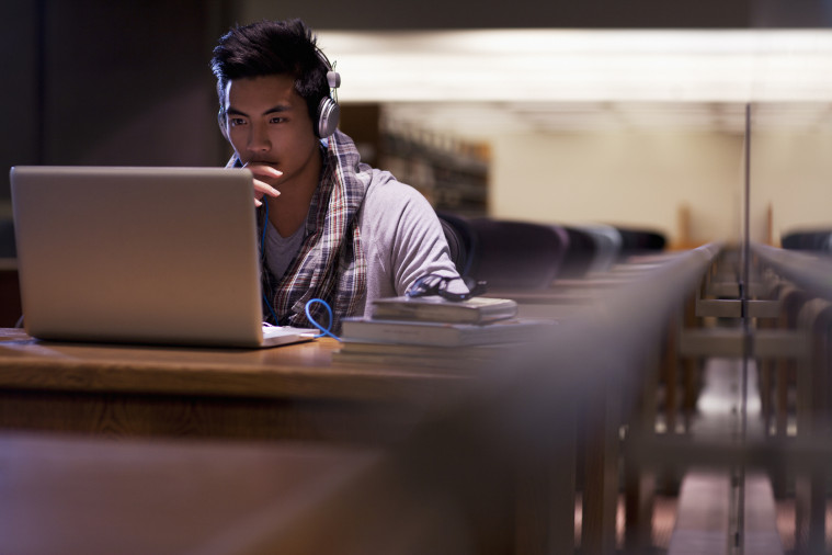 Un jeune étudiant portant un casque argenté blancs regarde son laptop de manière pensive. Il est dans une bibliothèque. Sur son bureau se trouve une pile de livre sur laquelle est placée une paire de lunettes.