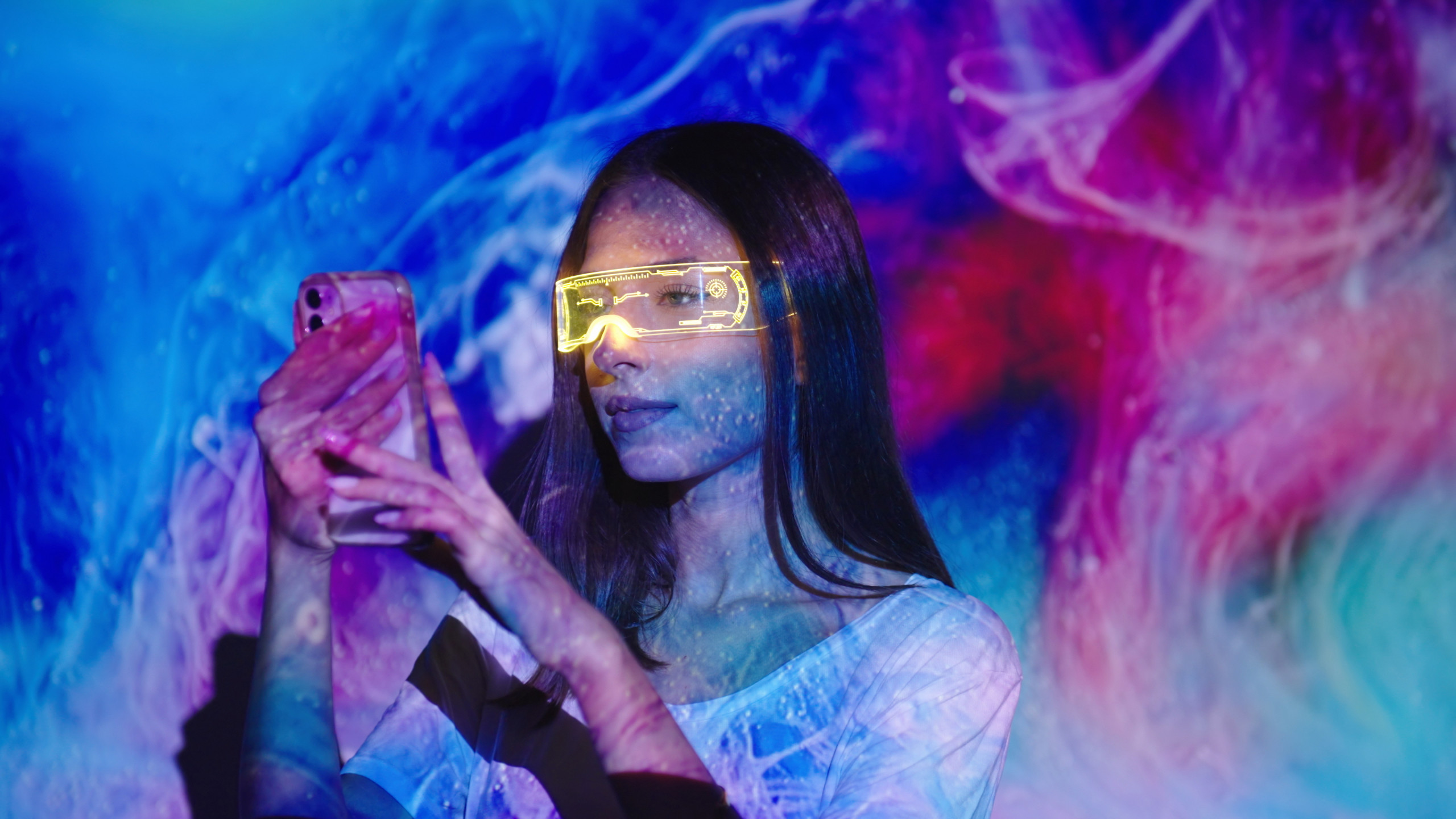 Jeune femme portant des lunettes de réalitée augmentée et tenant son smartphone à deux mains. Elle est baignée de la lumière d'une oeuvre d'art projetée sur le mur derrière elle.