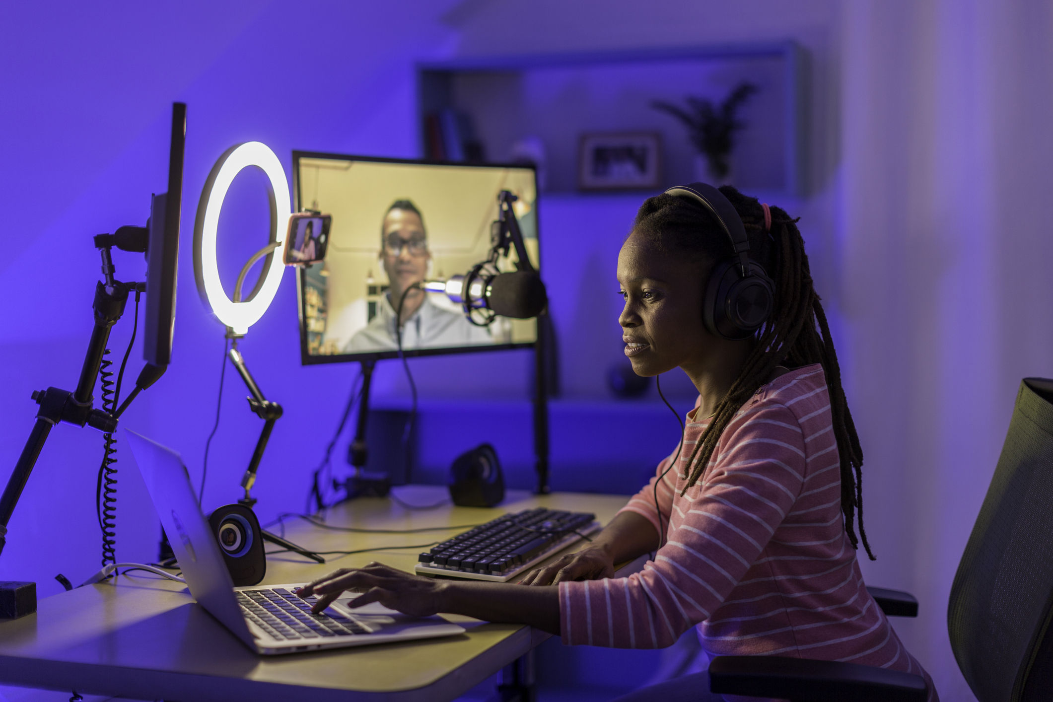 Jeune femme enregistrant une émission. Elle porte un casque, se tient devant un micro et un laptop, et est en vidéoconférence avec une jeune homme sur un écran fixe.