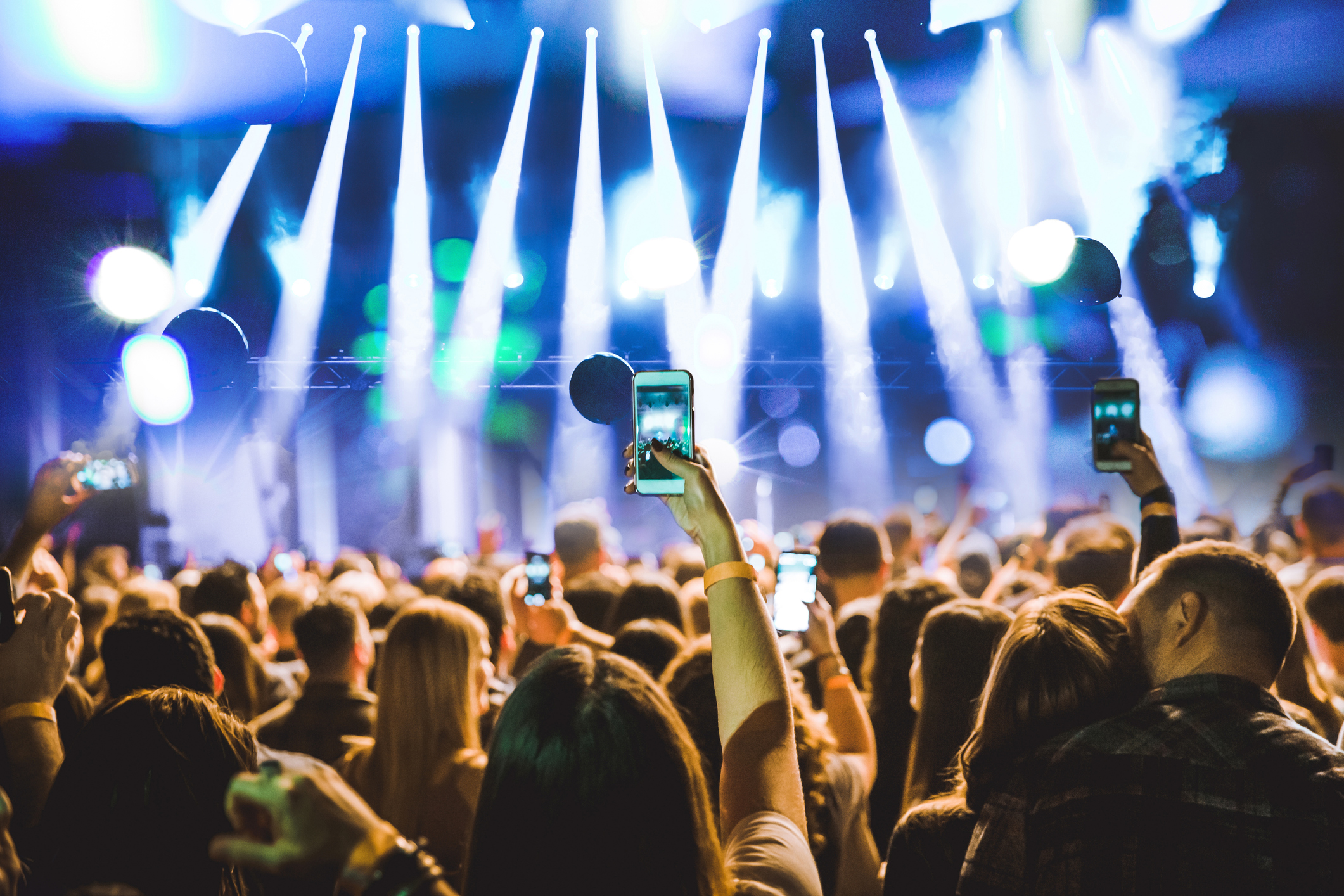 Une foule à un concert. Plusieurs personnes lèvent leurs téléphones pour filmer la scène, éclairé par des spots blancs. Le fond de l'image est bleu.