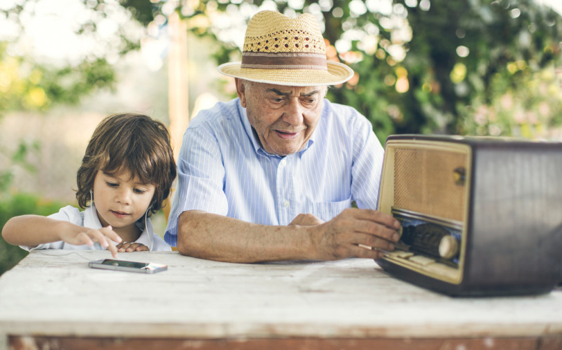 Un homme âgé portant un chapeau écoute la radio, pendant que son petit-fils portant des écouteurs écoute de la musique depuis un smartphone.