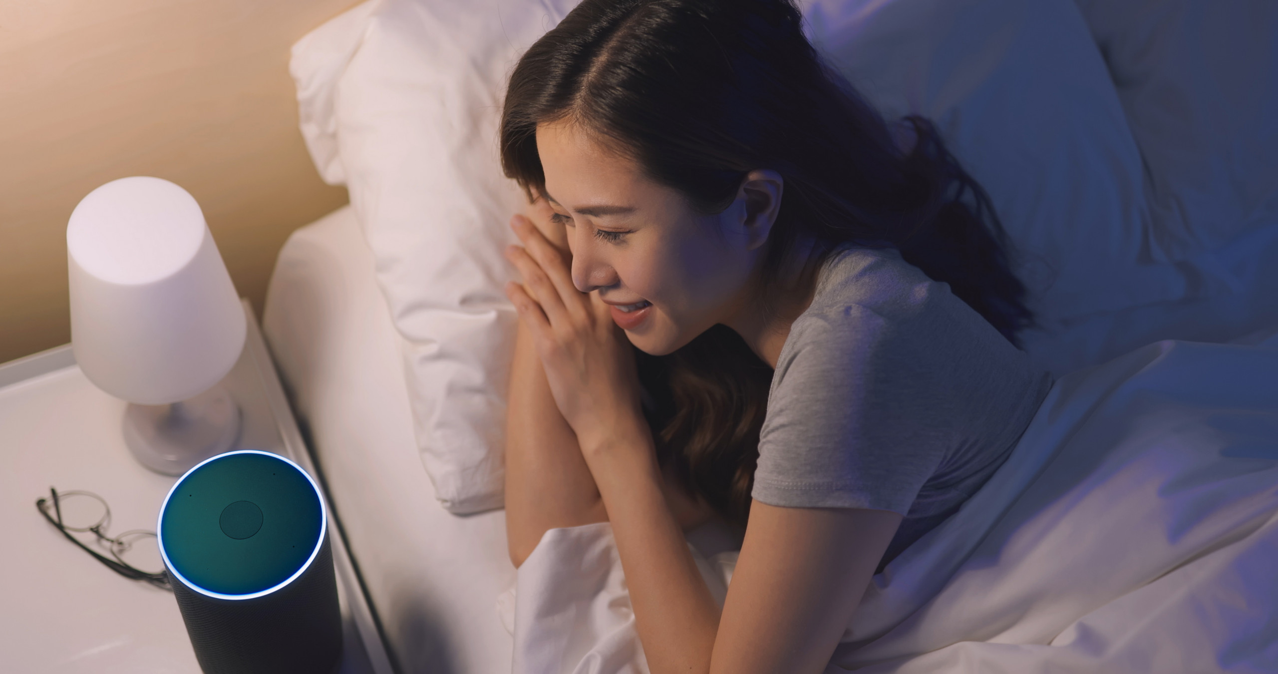 Une jeune femme asiatique allongée dans son lit parle à son enceinte connectée, posée sur sa table de chevet à côté de ses lunettes et d'une lampe.