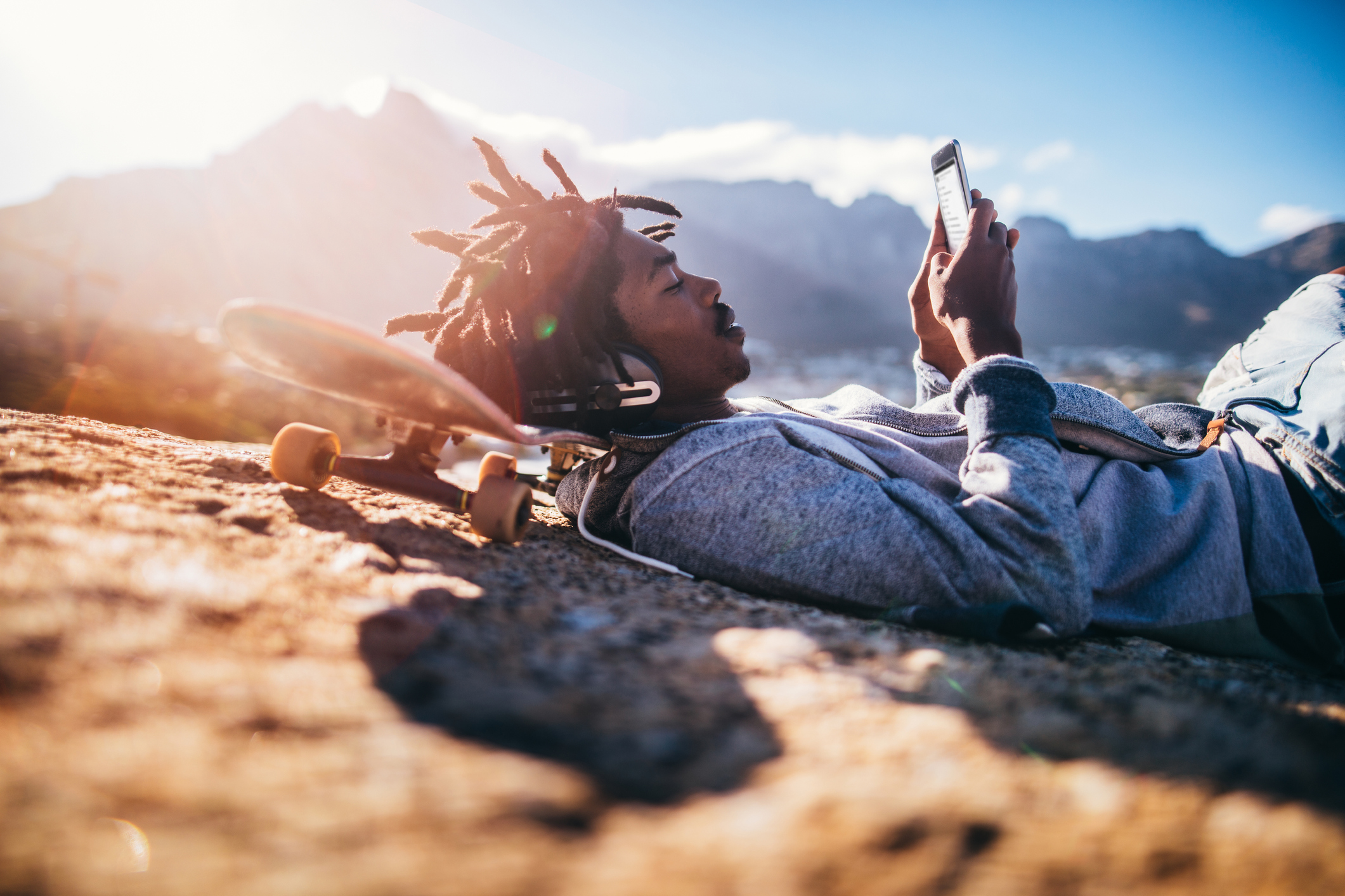 Un jeune homme noir, la tête posée sur un skateboard, regarde son smartphone en portant un casque. En arrière-plan, on aperçoit des montagnes et le soleil.